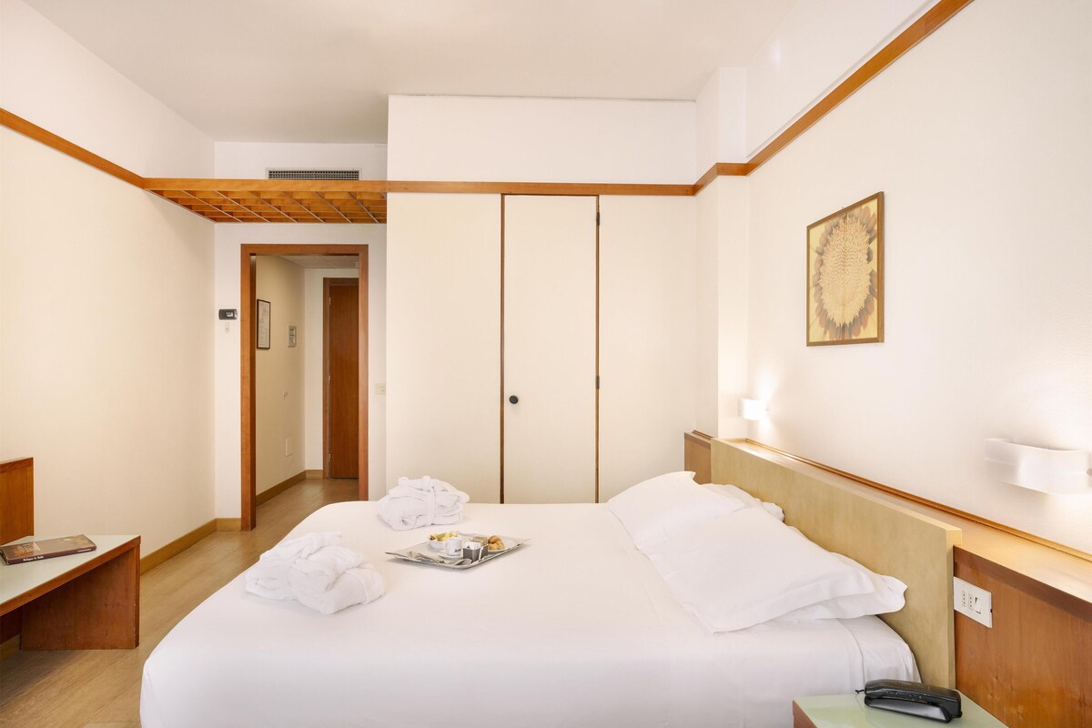 Une chambre d’hôtel avec un grand lit, une tête de lit en bois, une table de chevet avec une lampe et une porte donnant sur une autre pièce.