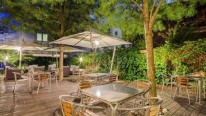 Eine Terrassensitzgruppe mit Tischen und Stühlen, umgeben von Pflanzen und Bäumen.
