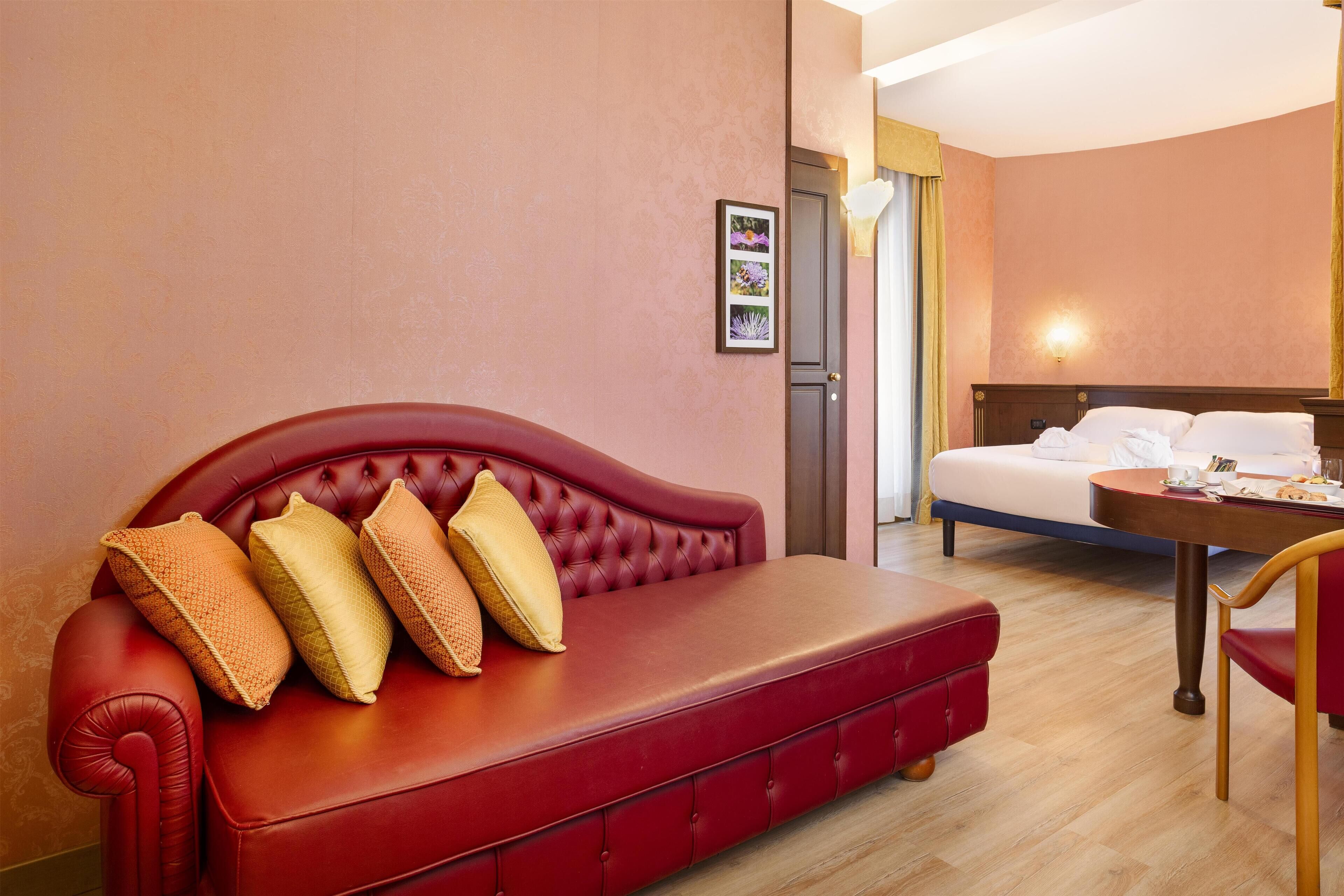 Een rode leren bank met vier kussens, een houten salontafel, een wit bed en een ingelijst beeld aan de muur.