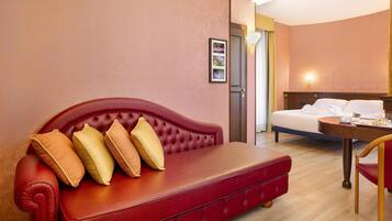 Een rode leren bank met vier kussens, een houten salontafel, een wit bed en een ingelijst beeld aan de muur.