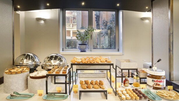 Ein Frühstücksbuffet mit verschiedenen Backwaren, einem Nutella-Glas und einer Pflanze am Fenster.