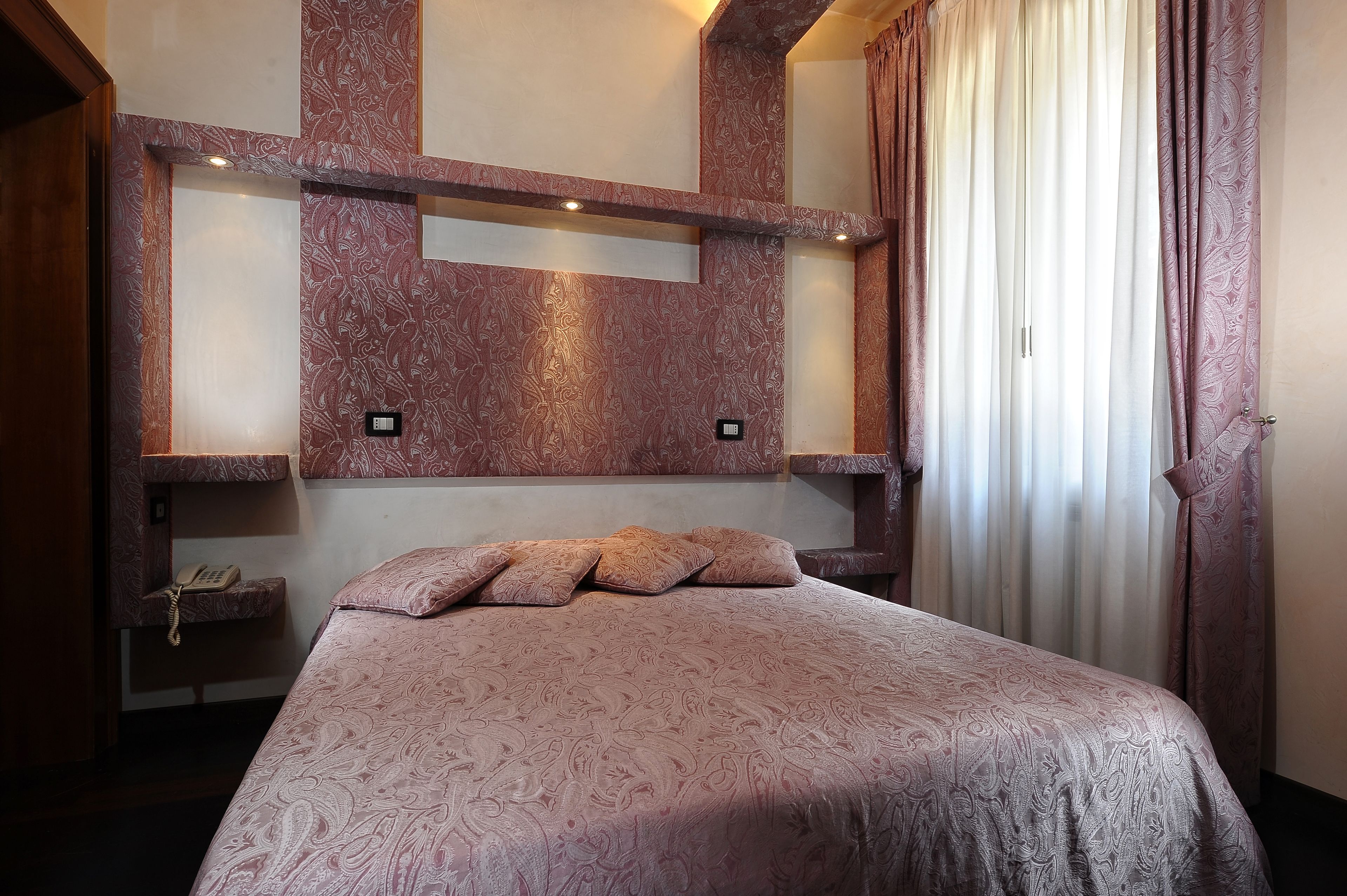 A hotel room with a large bed, a built-in shelf with a telephone, and a window with curtains.