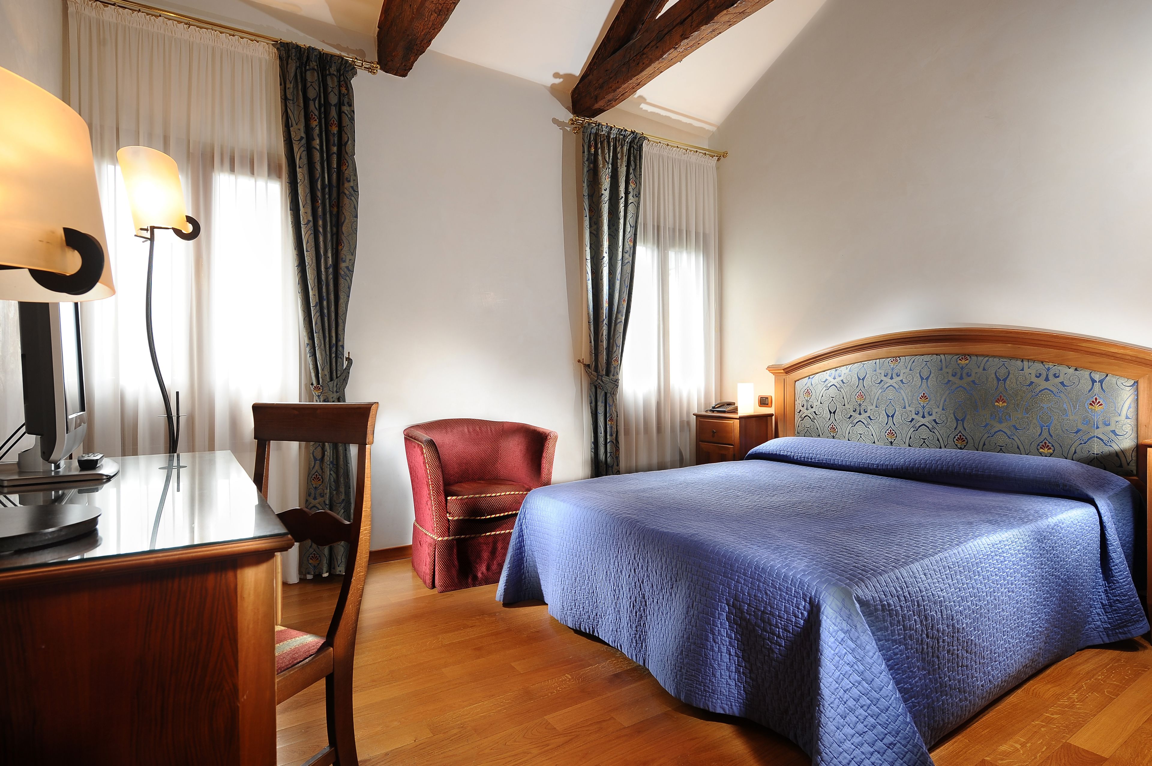 A hotel room with a wooden desk, a red chair, a bed with a blue quilt, and a patterned headboard.
