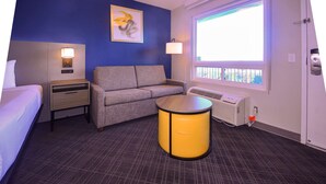 Une chambre d’hôtel avec un lit, un canapé, une petite table, une lampe, un tableau au mur et une fenêtre donnant sur un paysage.