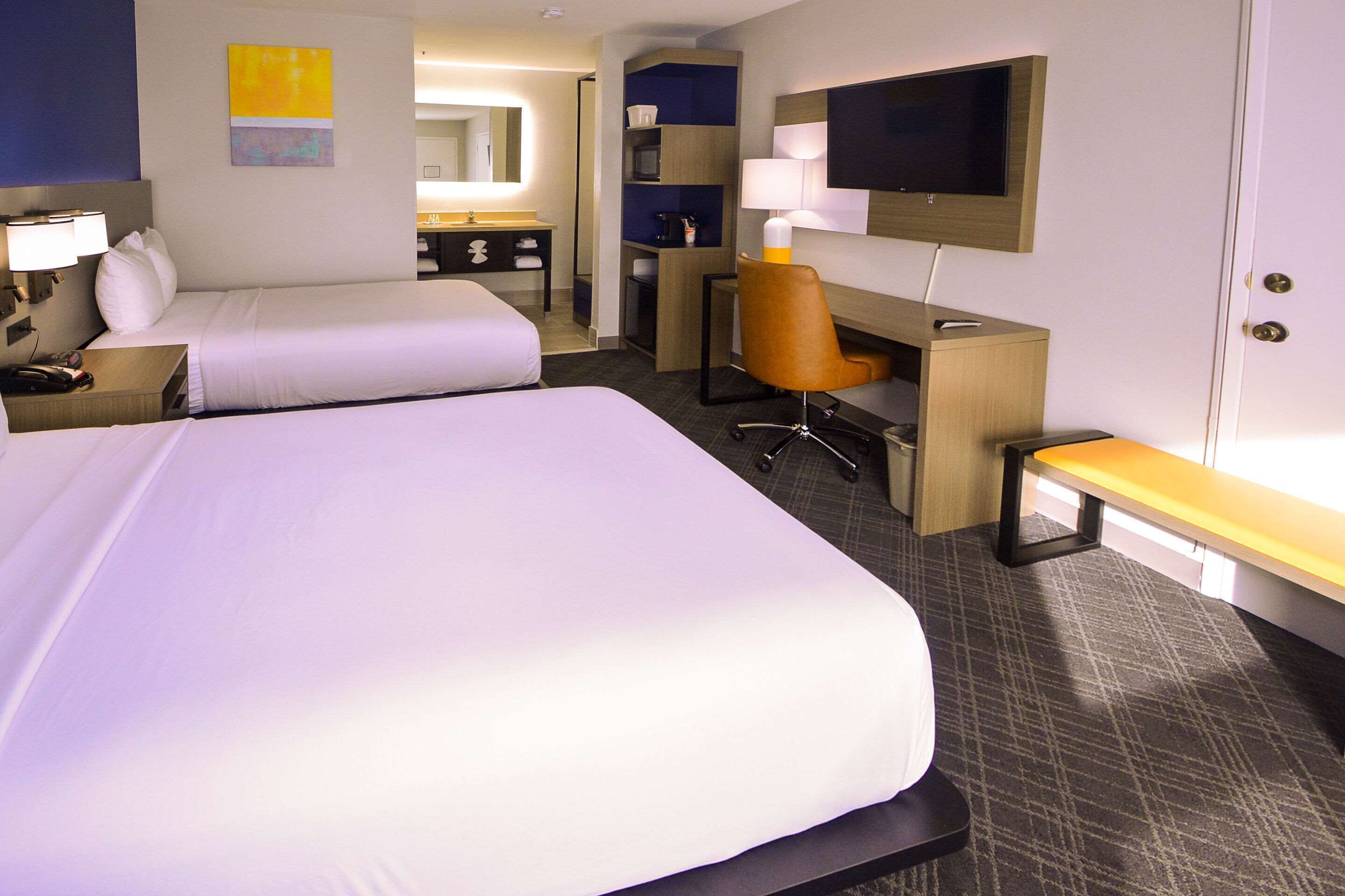 Quarto de hotel com duas camas, uma escrivaninha, uma cadeira, uma televisão e um banheiro visível ao fundo.
