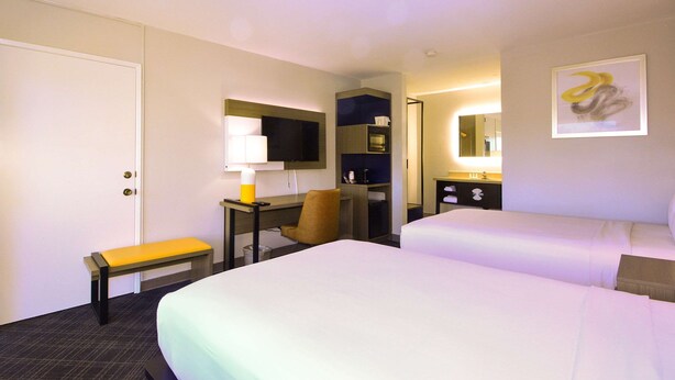 Une chambre d’hôtel comprenant un lit, un bureau avec une chaise, un banc, une télévision, un miroir et une salle de bain.