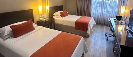 A hotel room with two beds, a desk, and a view of the city.