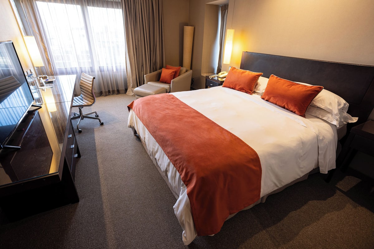 Ein Hotelzimmer mit einem großen Bett, einem Schreibtisch mit Computer, einem Stuhl und einer Lampe.