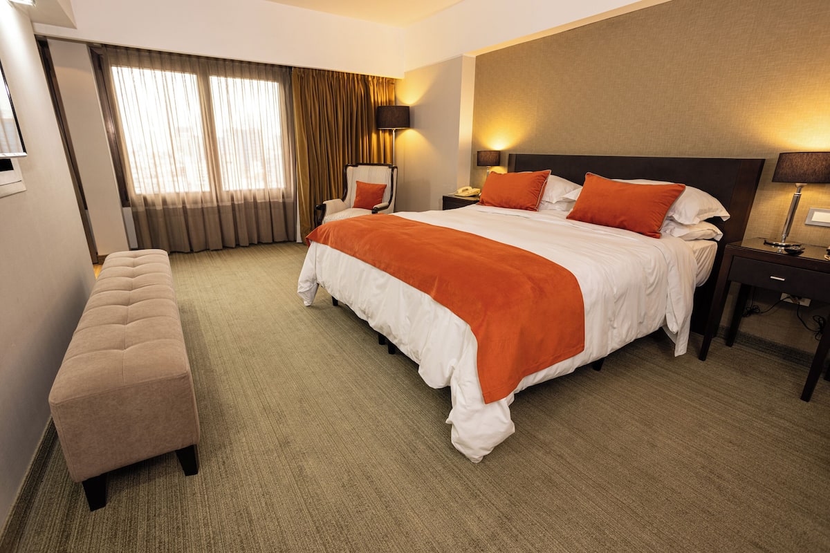 Ein Hotelzimmer mit einem großen Bett, einer Sitzbank, einem Schreibtisch und einem Fenster mit Vorhängen.