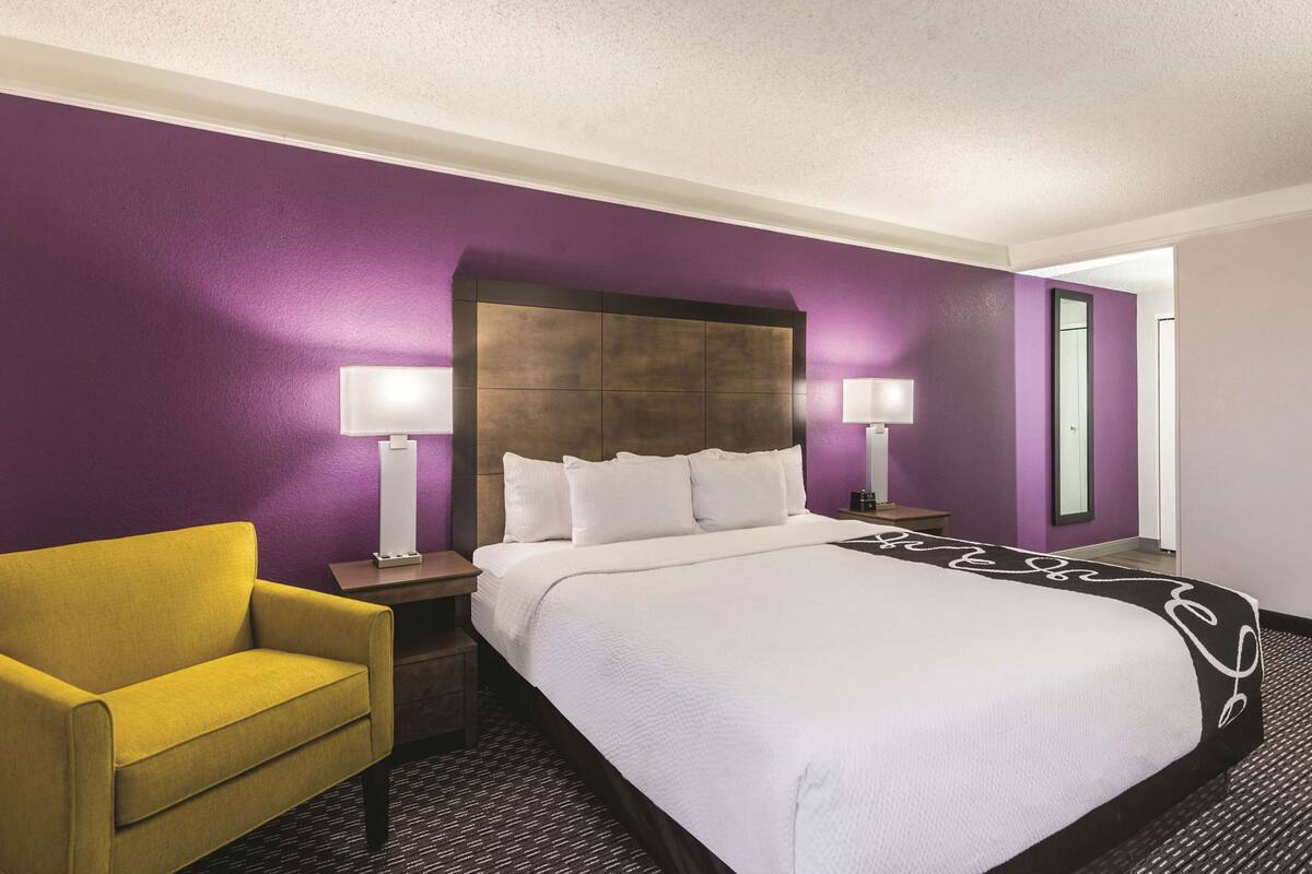 Una habitación de hotel con una cama grande, dos lámparas de noche, un sillón amarillo y paredes moradas.