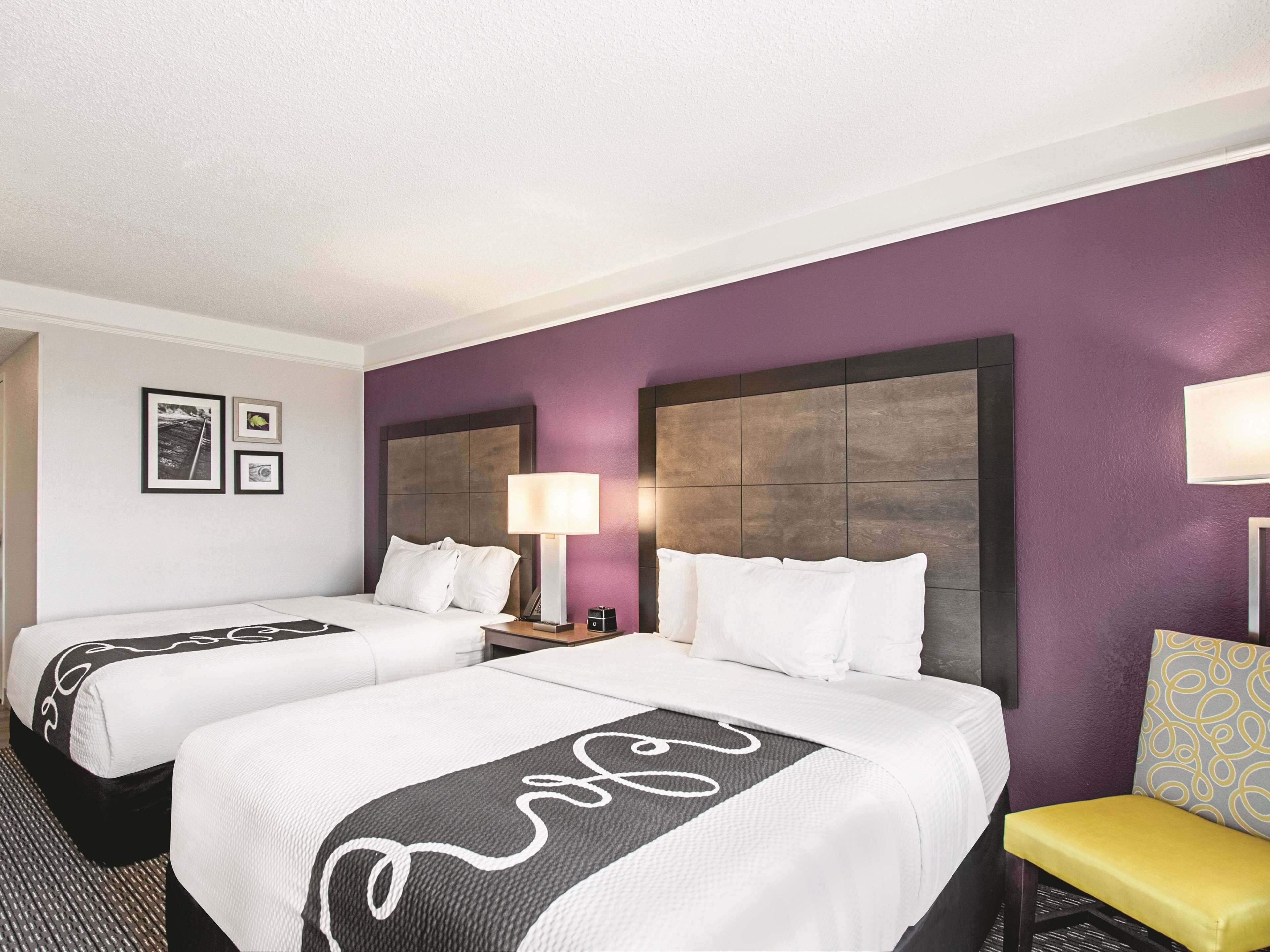 Habitación de hotel con dos camas, una mesita de noche, una lámpara, una silla y una pared con cuadros enmarcados.