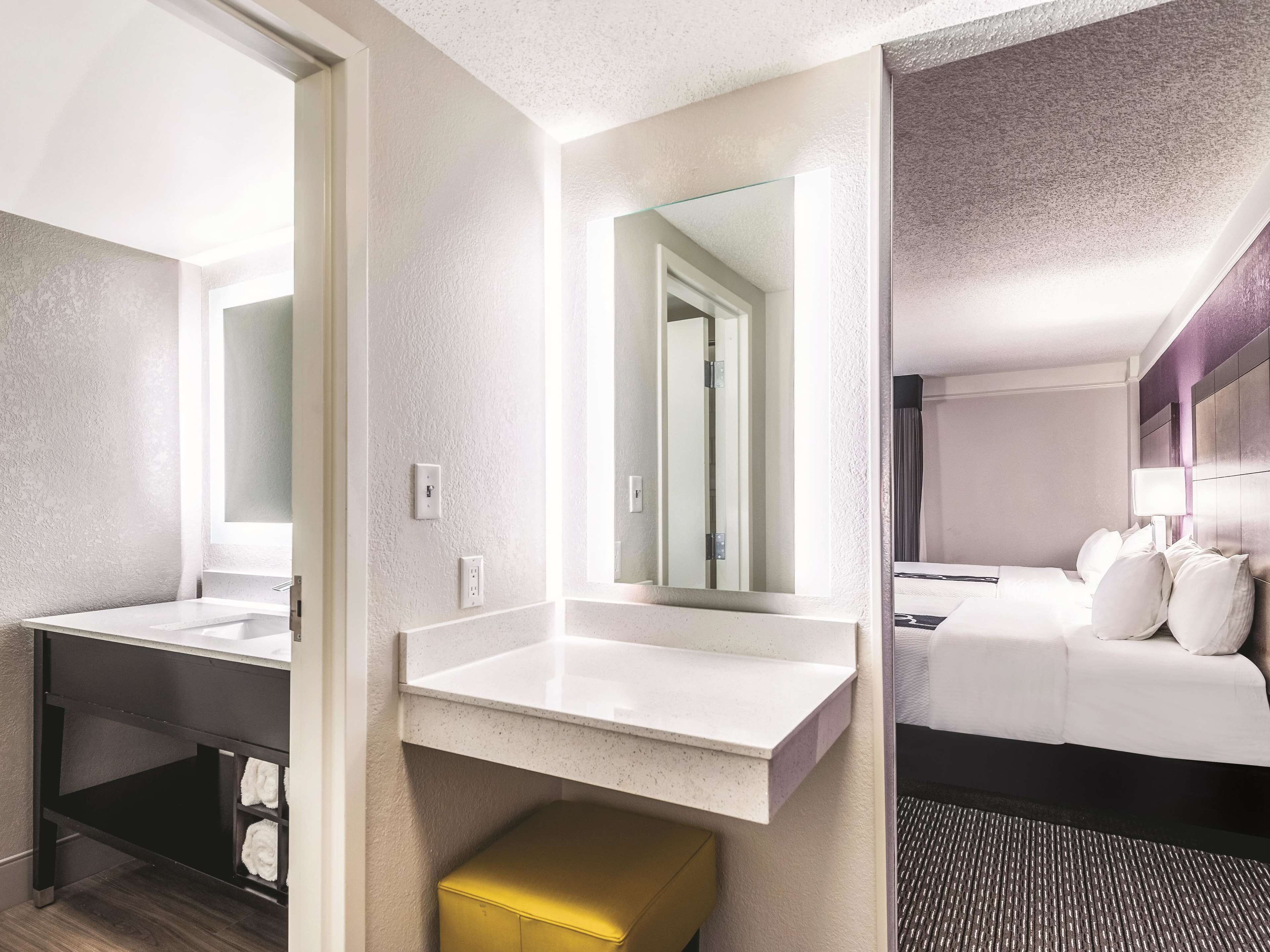 A hotel room with a large mirror, a white sink, a bed with white bedding, and a small yellow ottoman.