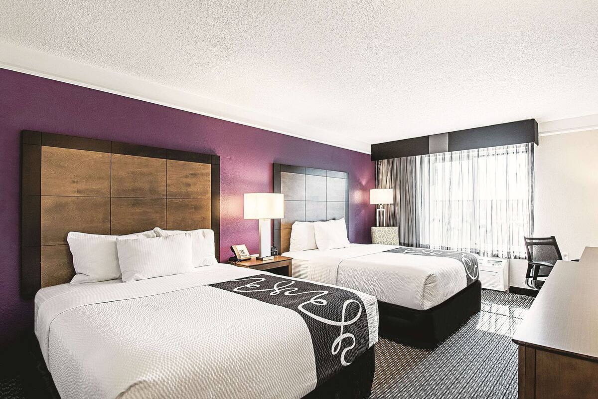 Una habitación de hotel con dos camas, un escritorio, una silla y un ventanal con cortinas.