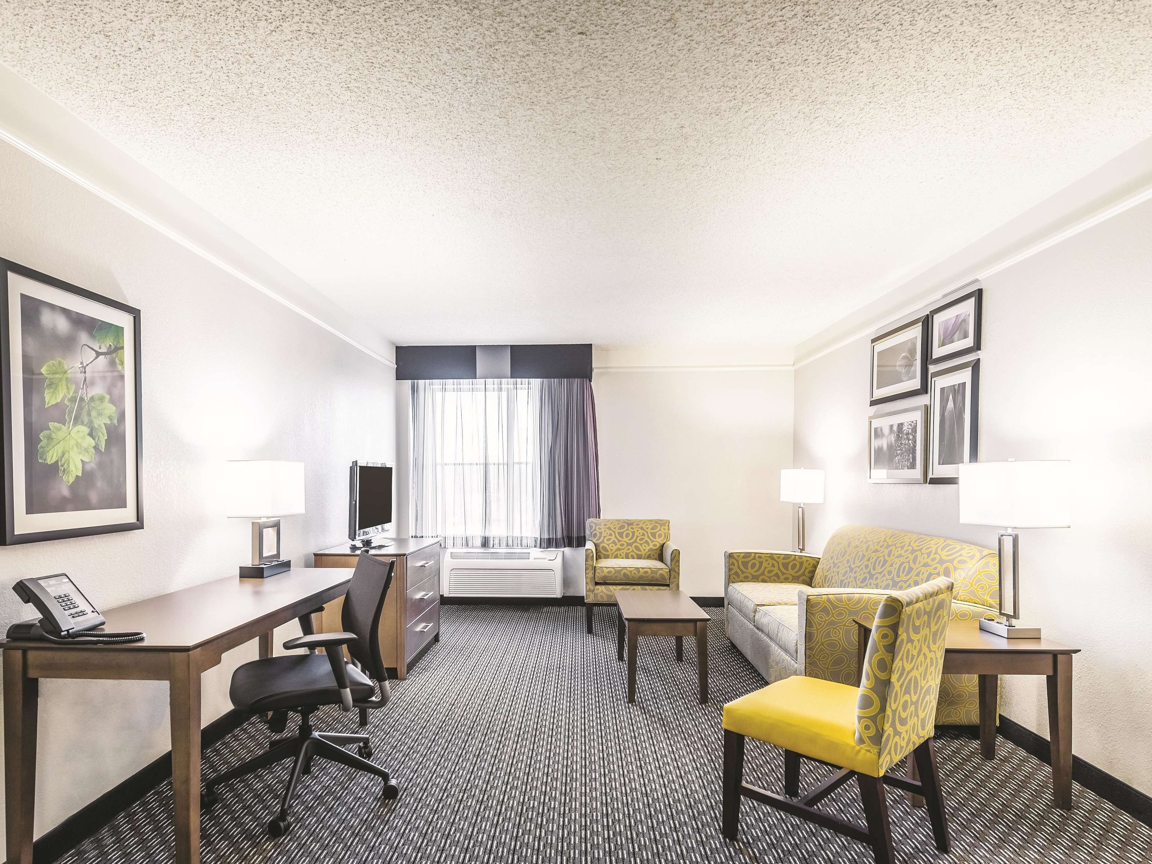 A hotel room with a desk, two chairs, a sofa, a TV, and a window with curtains.
