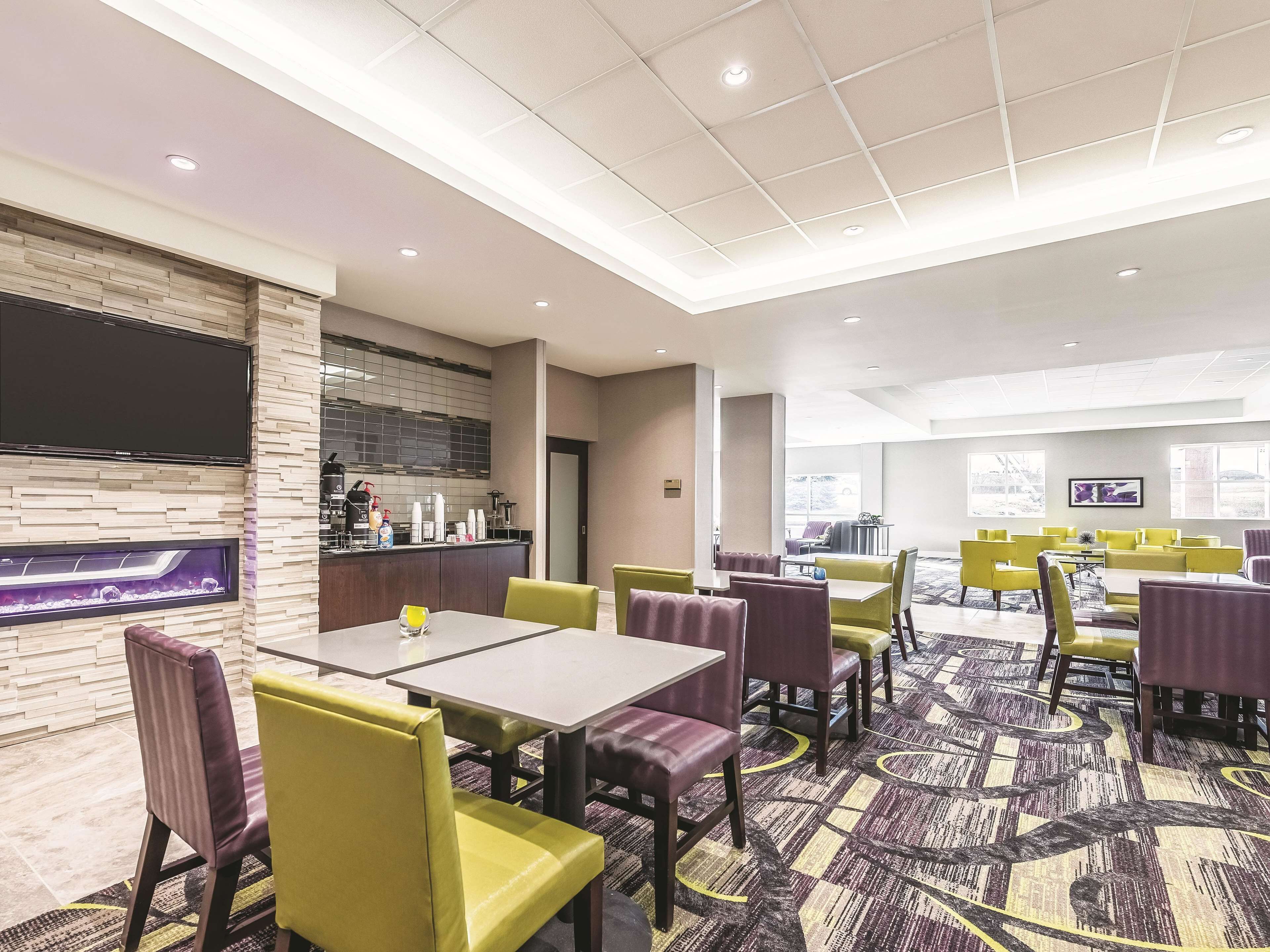 A hotel dining area with multiple tables and chairs, a kitchen area with a large screen, and a patterned carpet.