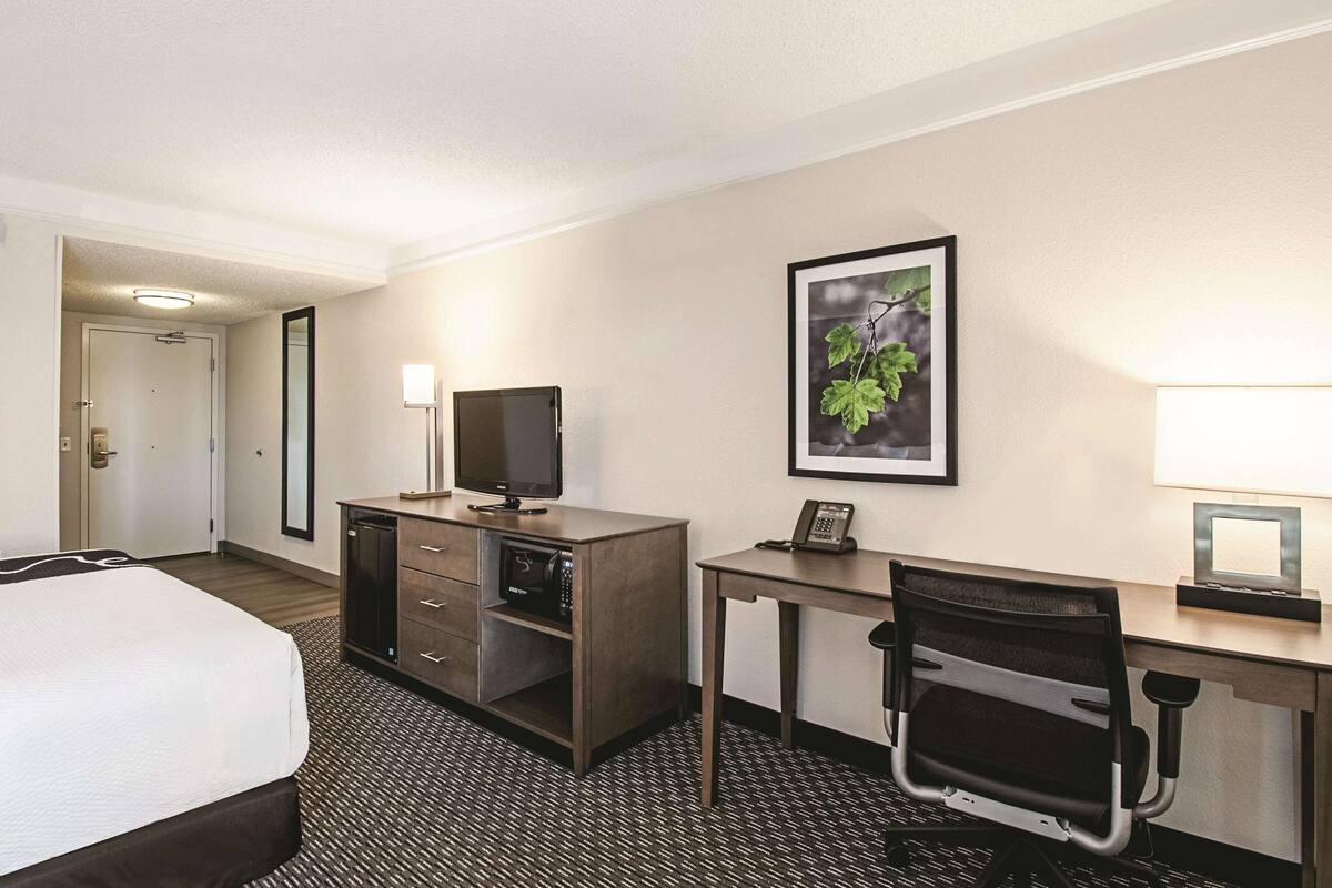 Habitación de hotel con cama, escritorio con computadora, silla, teléfono y un cuadro en la pared.