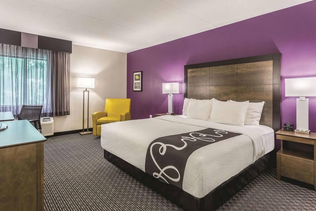 A hotel room with a large bed, a desk, a chair, and a nightstand with a lamp.