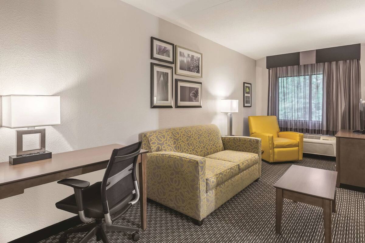 A hotel room with a sofa, a yellow armchair, a desk with a chair, a standing lamp, and framed pictures on the wall.