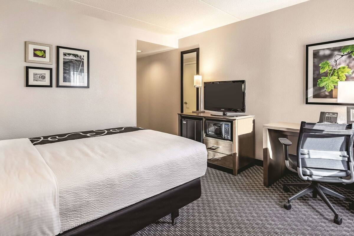 A hotel room with a bed, desk, chair, TV, and a small kitchenette.