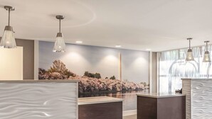 A reception area with a modern design, featuring a large wall mural of cherry blossoms and a calm water body. There are three reception desks with dark wood finishes and white textured panels. - La Quinta Inn & Suites by Wyndham Columbia / Fort Meade (Jessup)