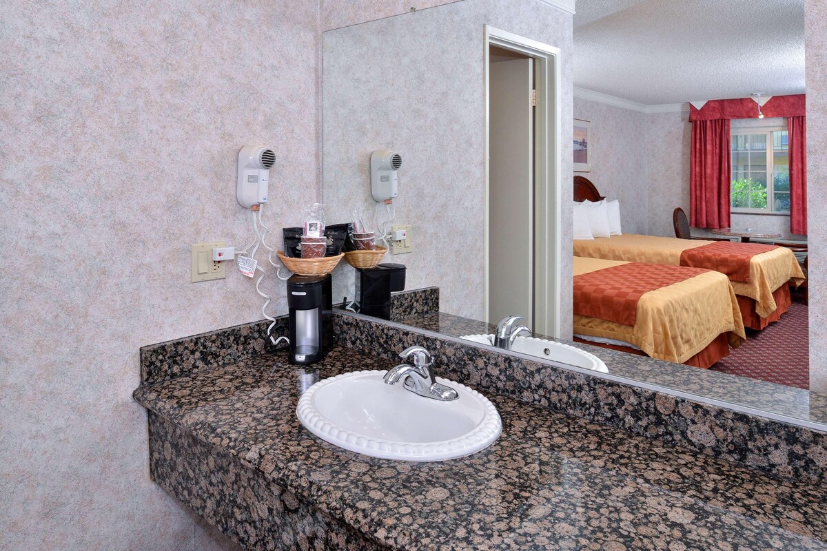 A hotel bathroom with a granite countertop sink, a mirror, two hair dryers, and a bed with red curtains.