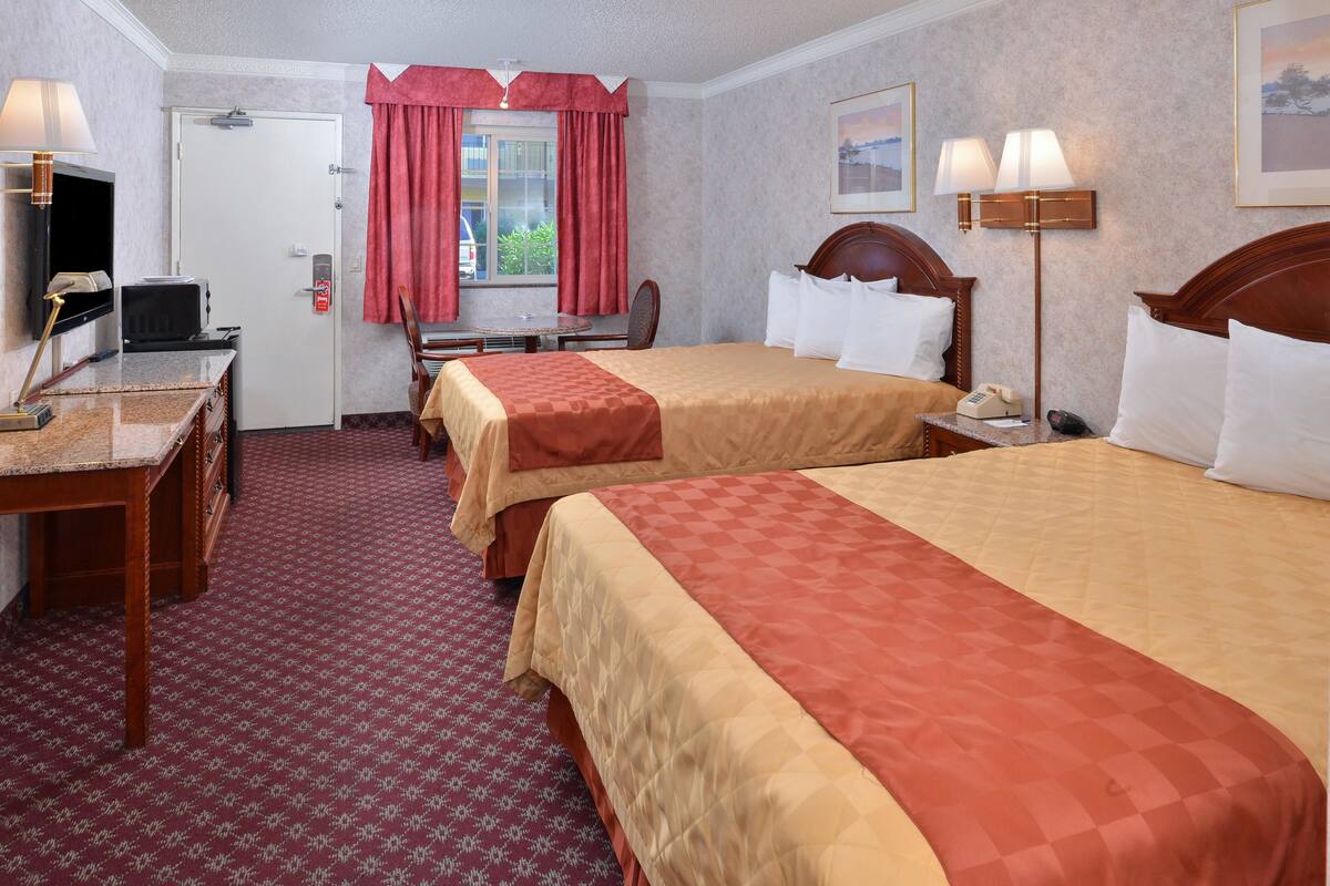 A hotel room with two beds, a desk with a computer, a telephone, and a window with red curtains.