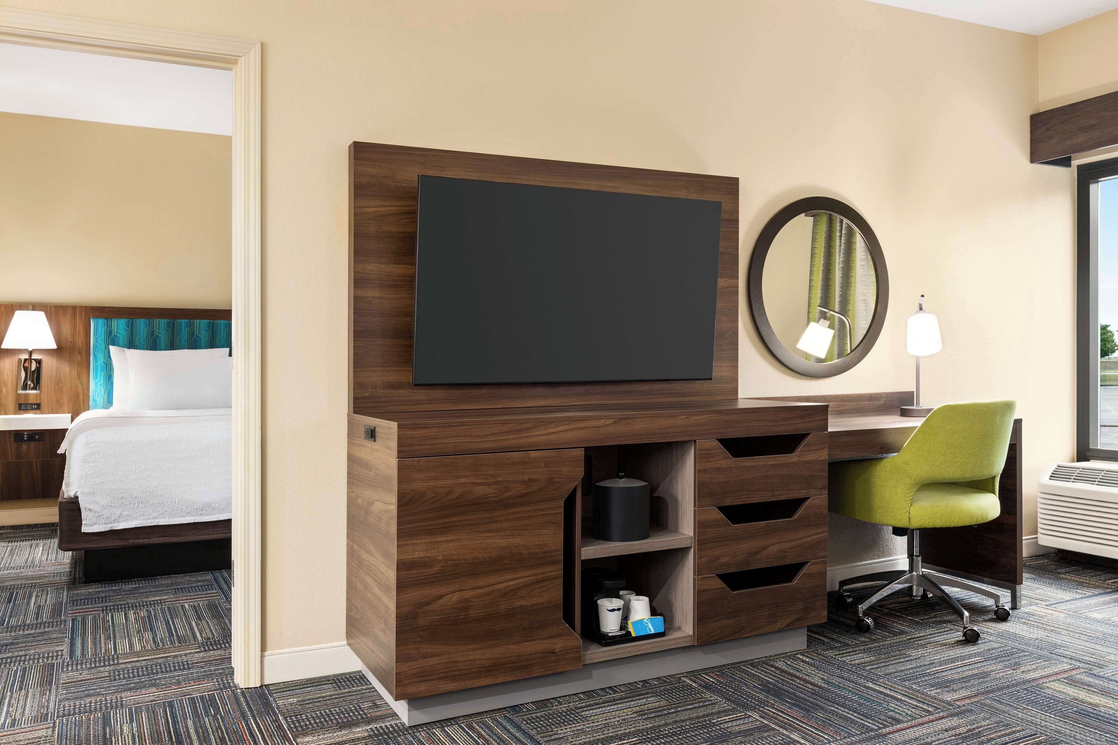 A hotel room with a bed, a desk with a chair, a flat-screen TV, a mirror, and a built-in heater.