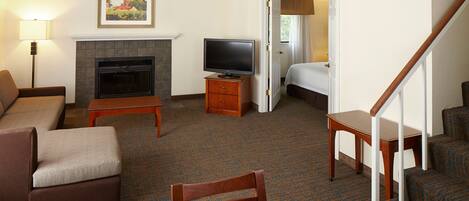 A hotel room with a sofa, a television on a wooden stand, a fireplace, a dining table, and a staircase.