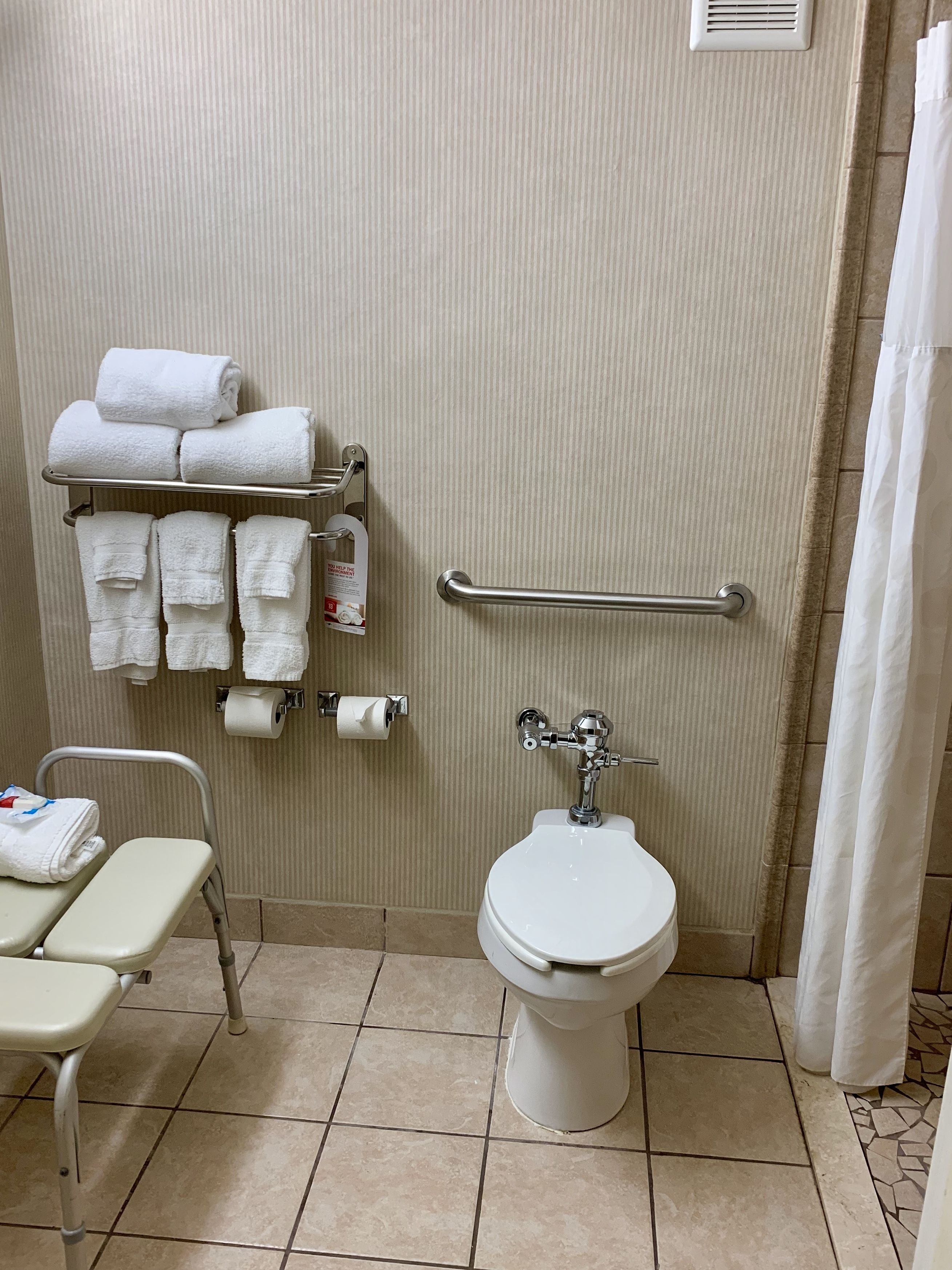 A bathroom with a toilet, a handrail, a towel rack with towels, a toilet paper holder, and a wheelchair-accessible bench.