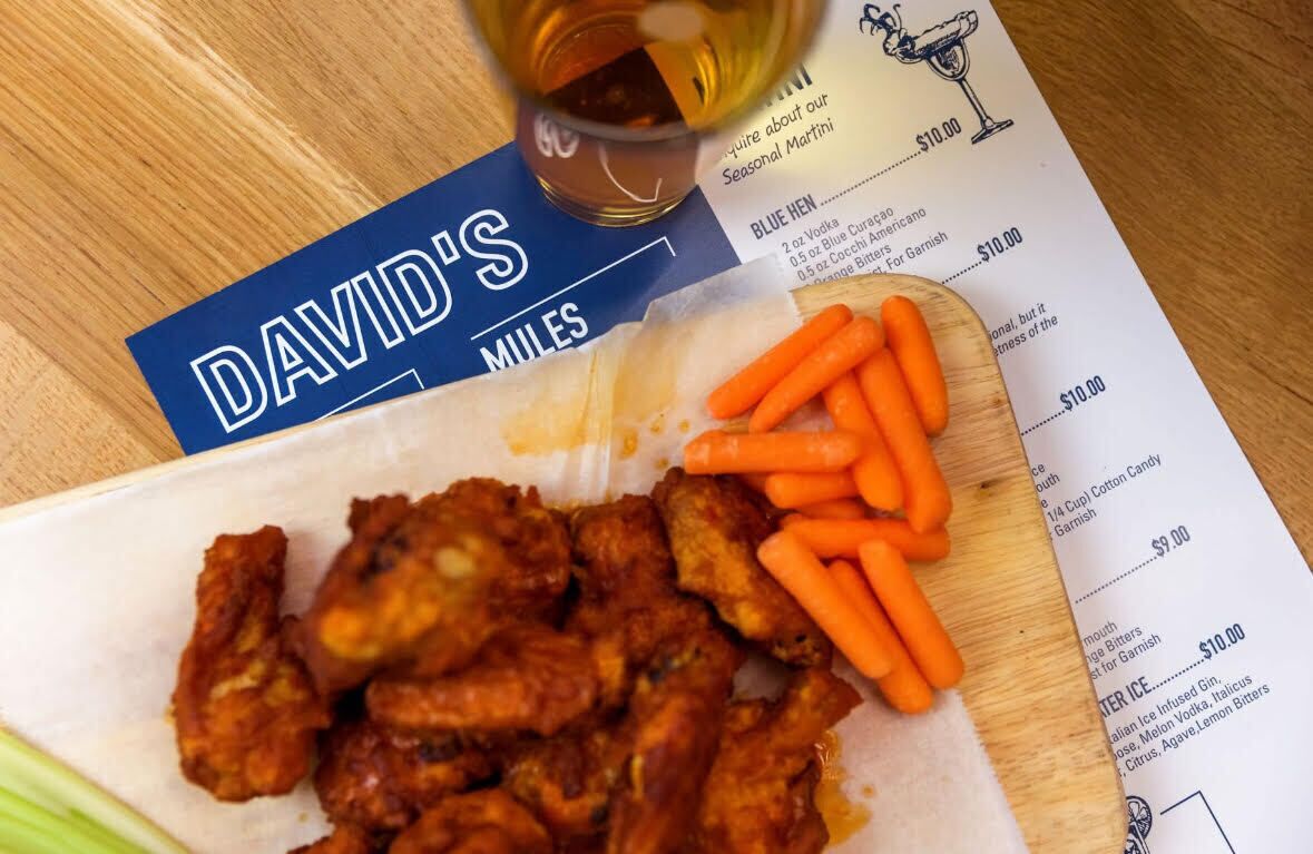 A menu with food items and prices, a bottle of beer, and a plate of chicken wings with carrots.