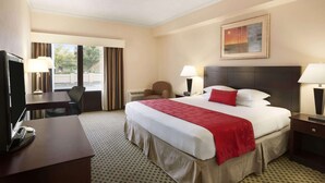 A hotel room with a large bed, a desk, a chair, a TV, and a window with curtains. - Ramada by Wyndham Newark/Wilmington (Newark)