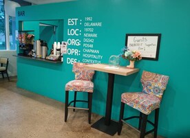 Una zona de bar color turquesa con un cartel que indica año de fundación, ubicación, organización y diseño. Dos sillas con motivos decorativos están dispuestas alrededor de una mesa con un florero.