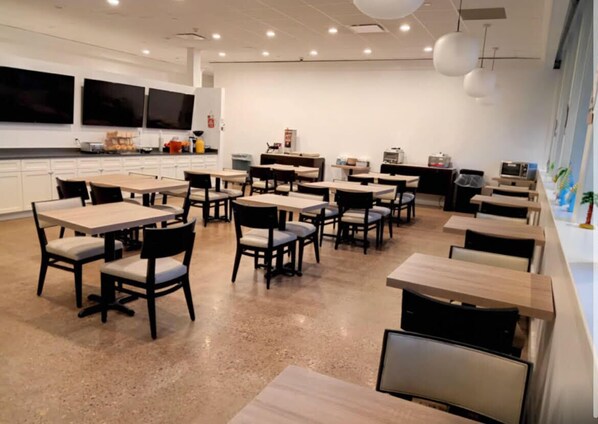 A modern dining area with multiple tables and chairs, a kitchen area with appliances, and a large window providing natural light. - Ramada by Wyndham Newark/Wilmington (Newark)