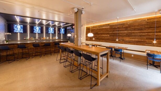 A modern bar area with a long wooden bar counter, multiple bar stools, and a seating area with tables and chairs. - Ramada by Wyndham Newark/Wilmington (Newark)