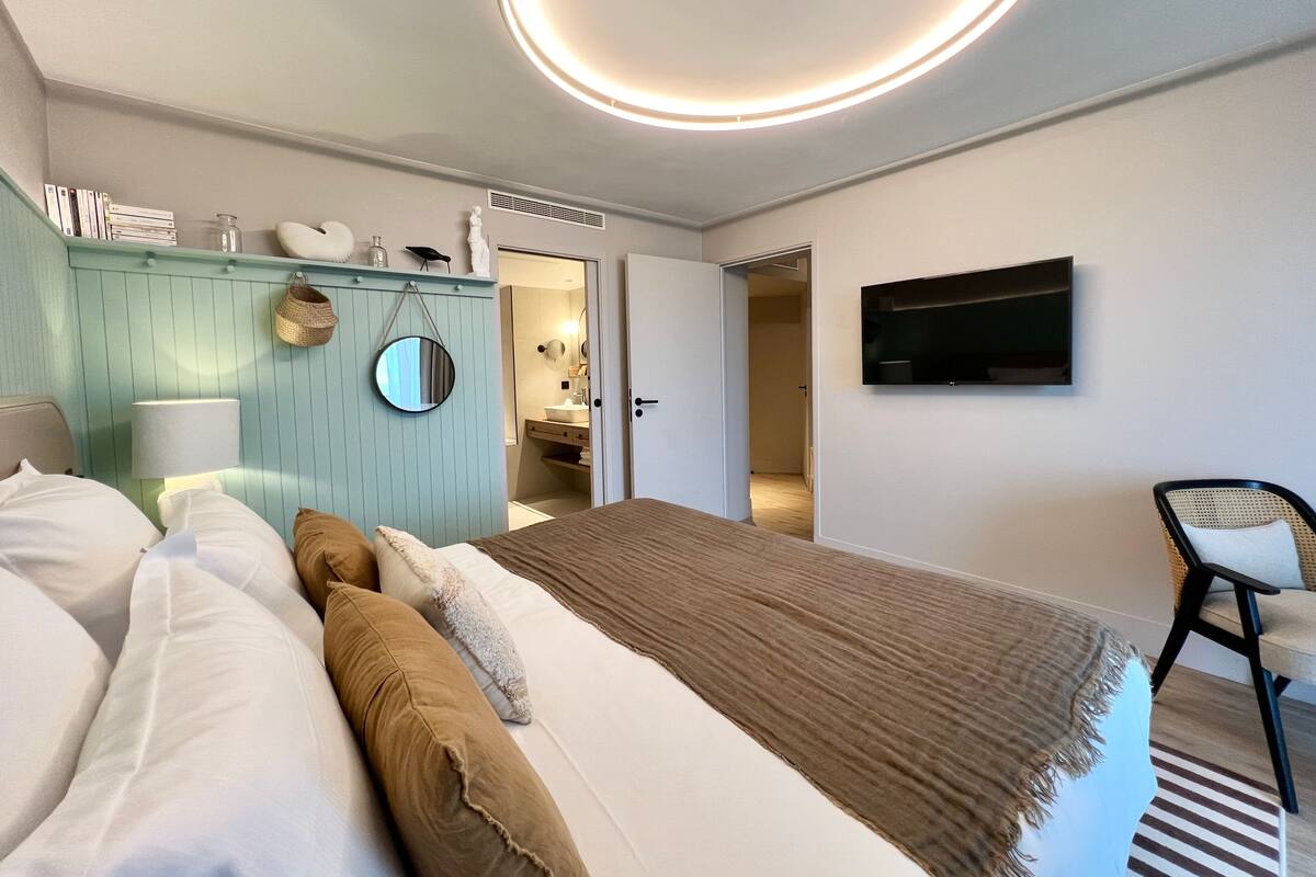 Une chambre moderne avec un lit, une télévision, un miroir et une salle de bain visible à travers une porte ouverte.