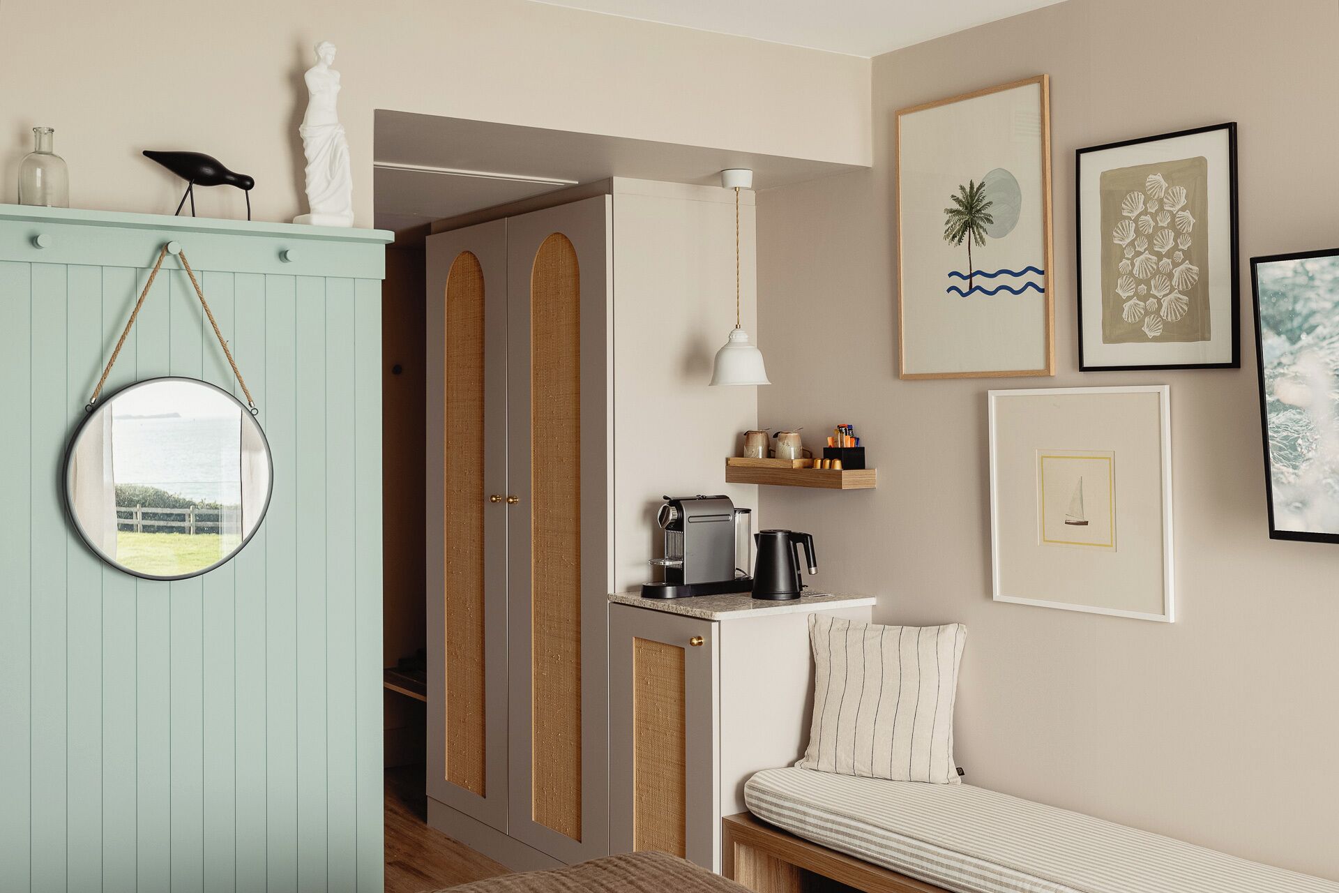 A room with a light blue wardrobe, a round mirror, a bed with striped bedding, a small coffee table, and framed artwork on the wall.