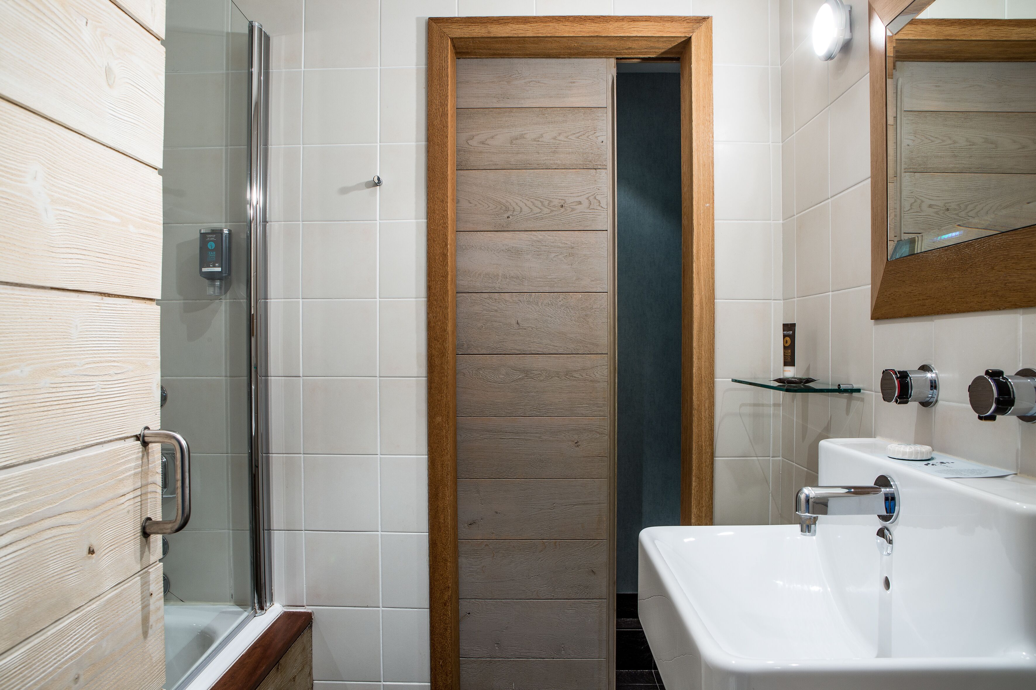 A modern bathroom with a glass shower enclosure, a wooden door, a white sink, and a mirror.