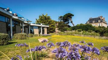 Ein modernes Gebäude mit Glasfassade, ein traditionelles Haus mit dunklem Dach und ein gepflegter Garten mit violetten Blumen.