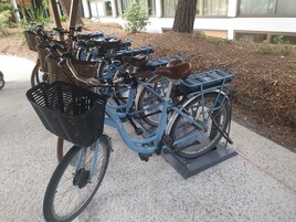 A blue bicycle with a basket and a kickstand, parked on a concrete surface.
