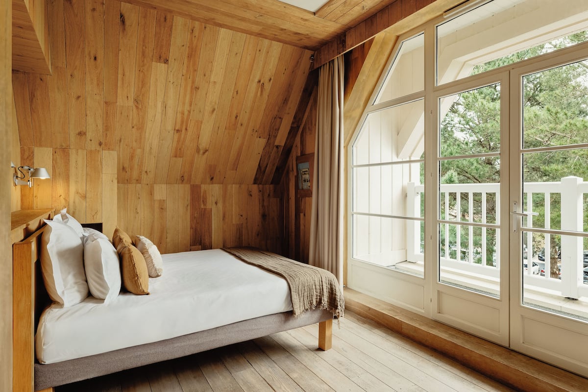 Une chambre avec un plafond en bois, un lit avec des oreillers, une fenêtre donnant sur des arbres et une clôture blanche à l’extérieur.