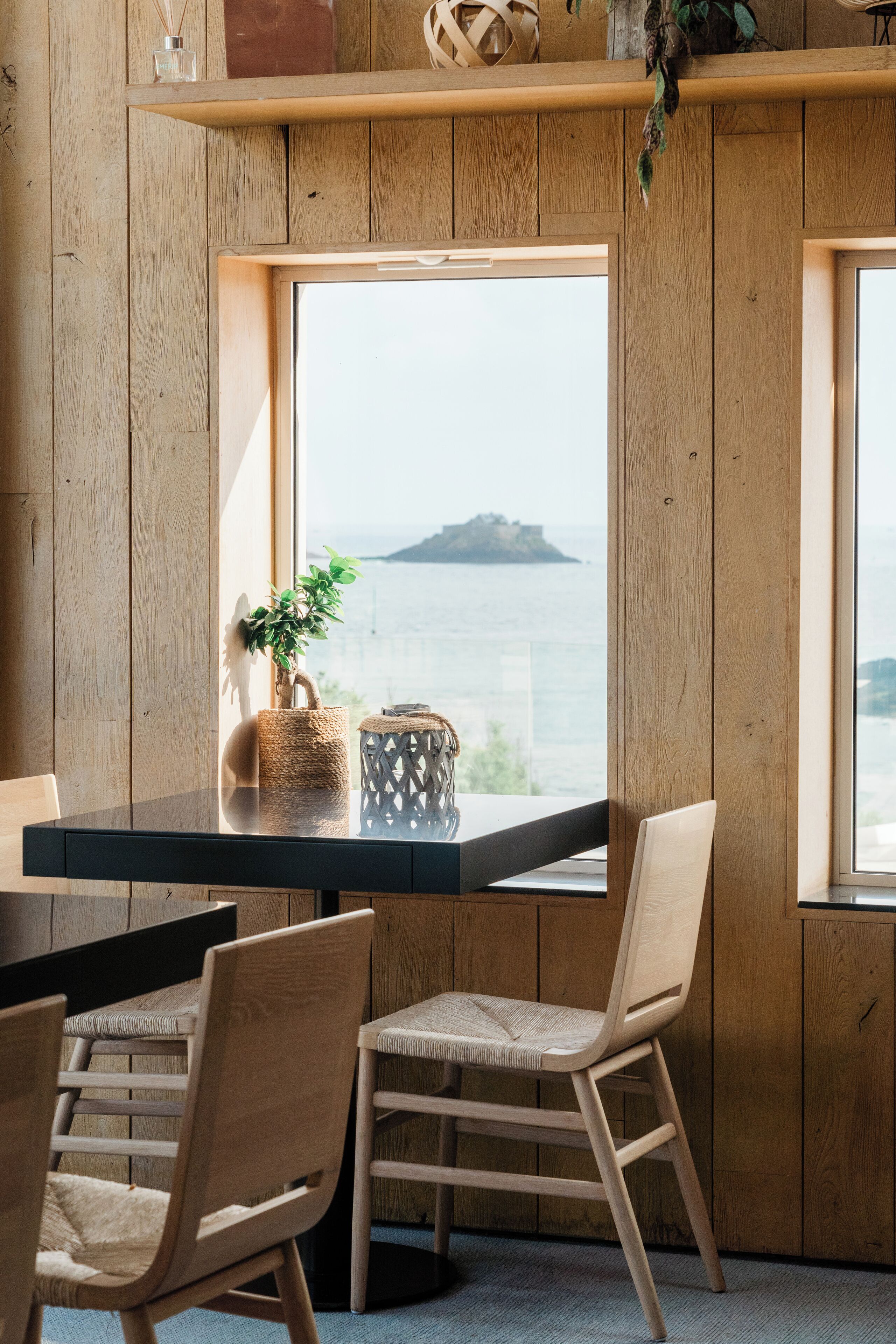 A wooden interior with a view of the sea, a table with chairs, and decorative items on a shelf.