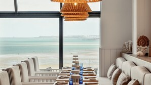 Ein Essbereich mit Blick auf den Strand, ausgestattet mit einem langen Tisch, blauem Glas und Geschirr, einer gewobenen Pendelleuchte und einer Couch mit Kissen.