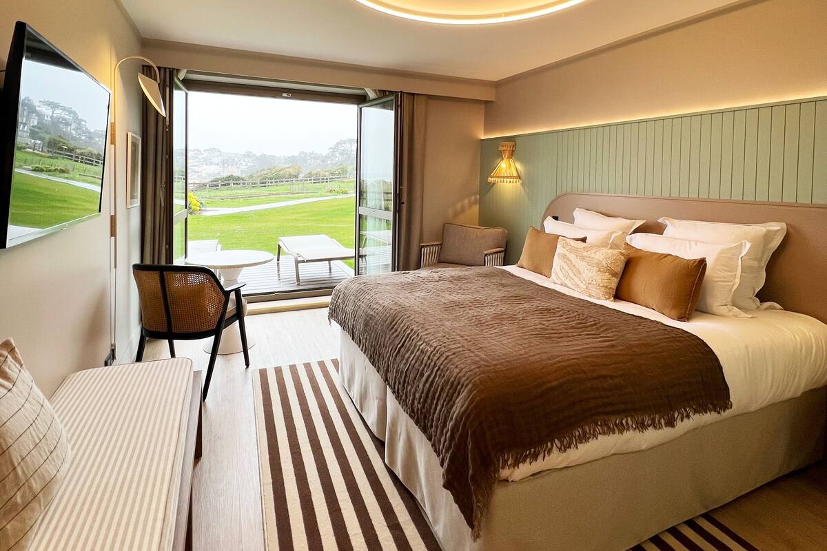 Une chambre d’hôtel moderne dotée d’un grand lit, d’un canapé, d’un fauteuil et offrant une vue sur un terrain de golf.