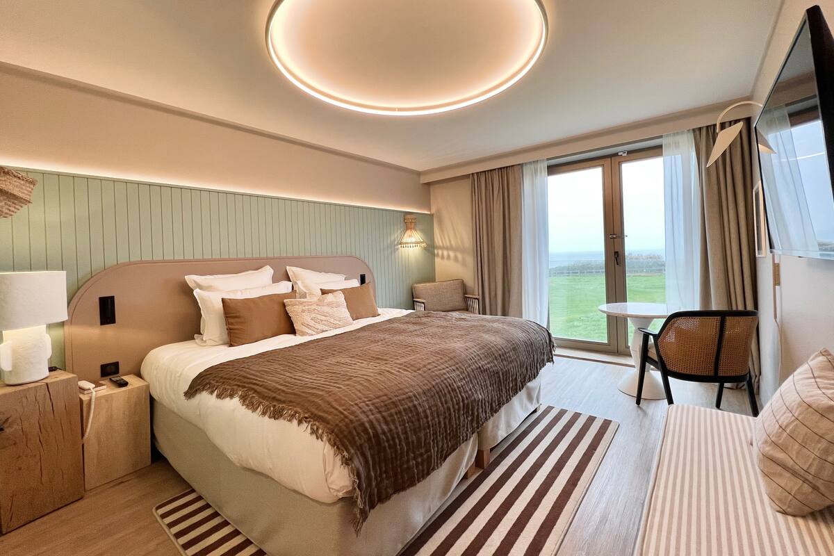 Une chambre d’hôtel moderne avec un grand lit, une table de chevet avec une lampe, une chaise et une vue sur l’extérieur.