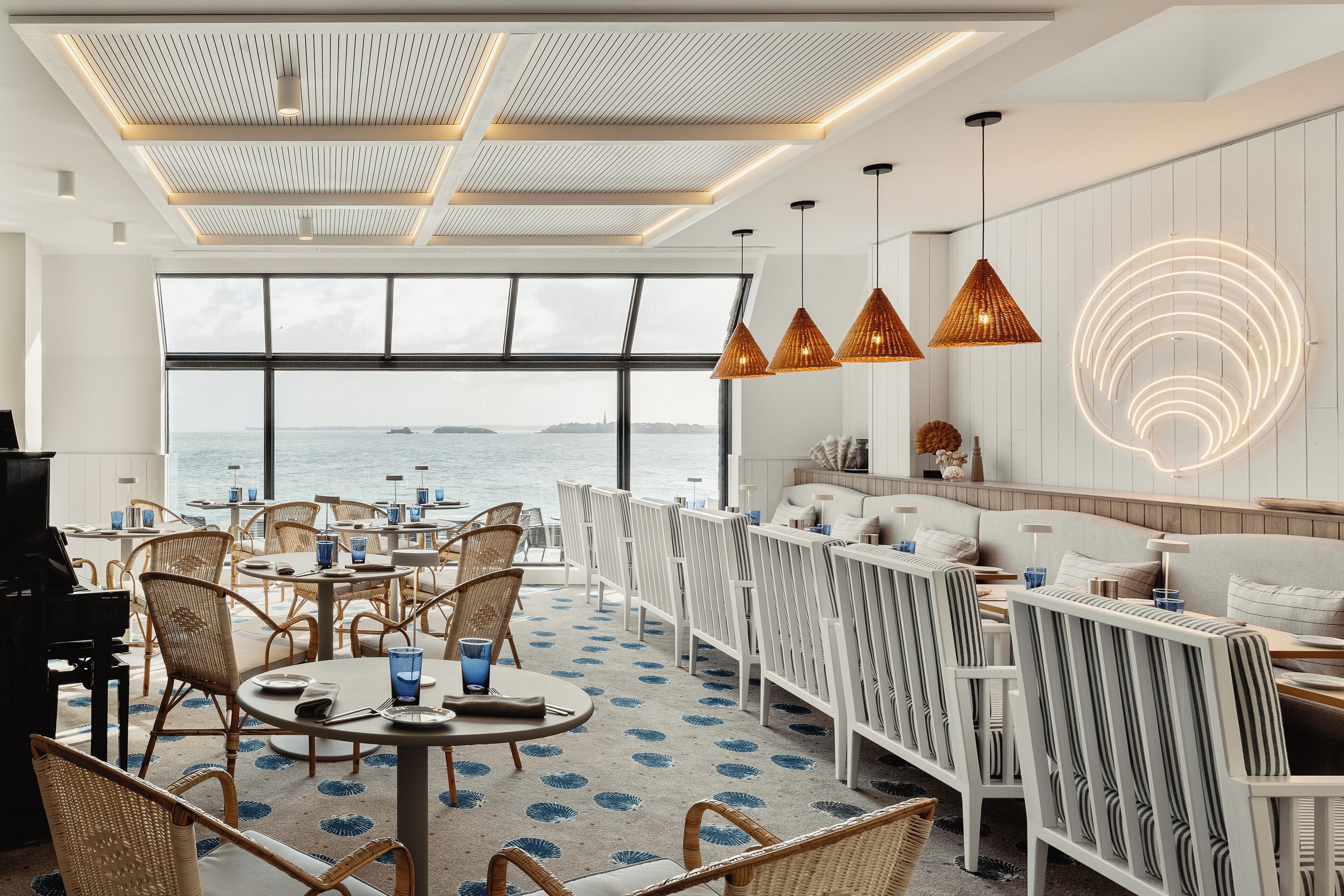 Un restaurant moderne offrant une vue sur la mer, avec des meubles en osier, des luminaires suspendus et un sol à motifs.