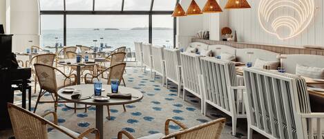Un restaurant moderne offrant une vue sur la mer, avec des meubles en osier, des luminaires suspendus et un sol à motifs.
