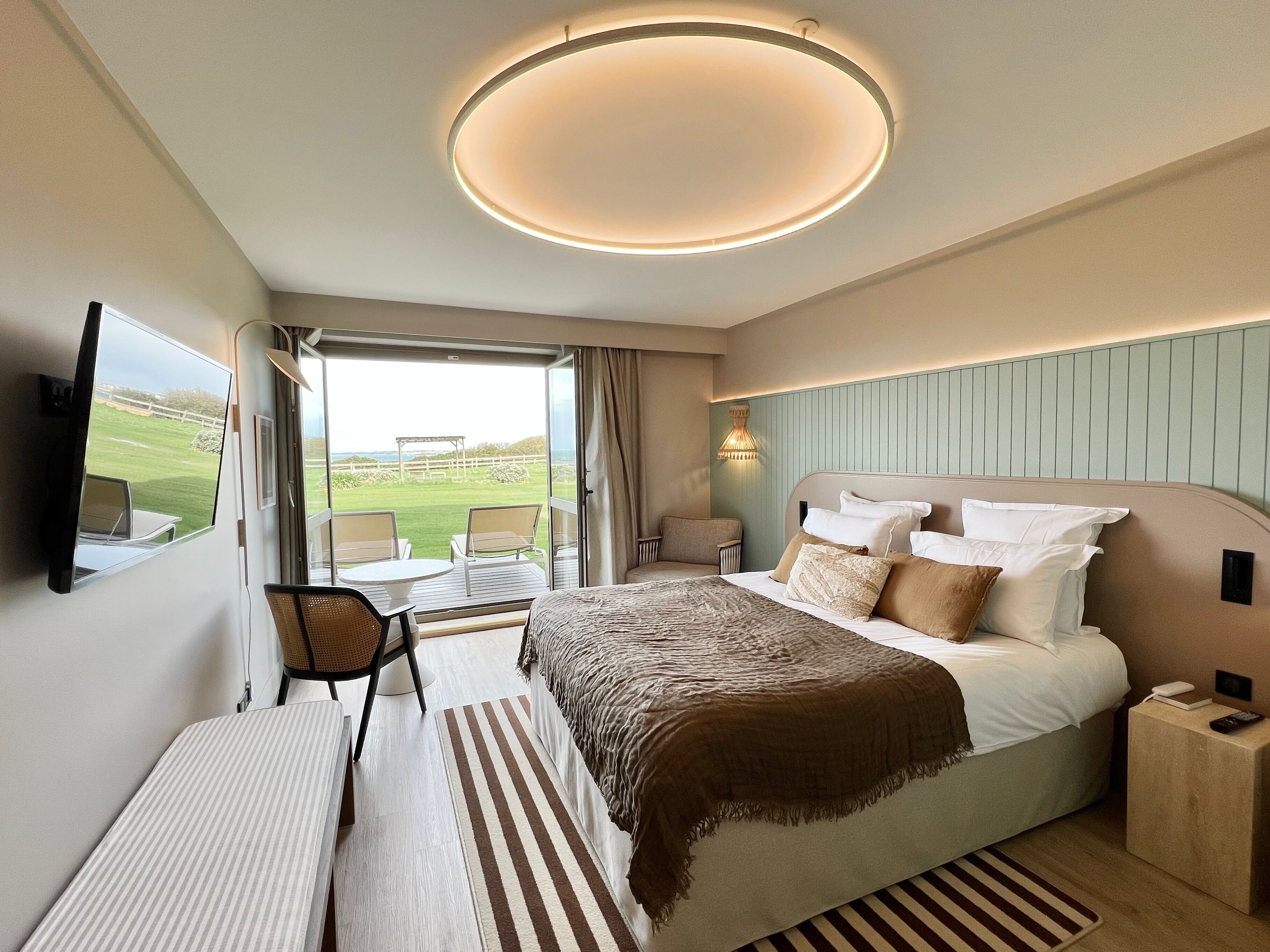 A hotel room with a large bed, a desk, a chair, a TV, and a view of a green landscape.
