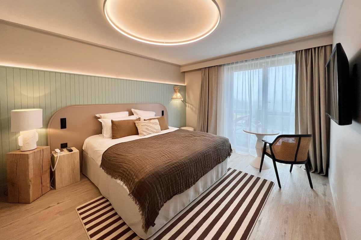 Une chambre d’hôtel moderne dotée d’un grand lit, de tables de chevet, d’un téléviseur à écran plat, d’une petite table avec une chaise et d’une grande fenêtre avec des rideaux.