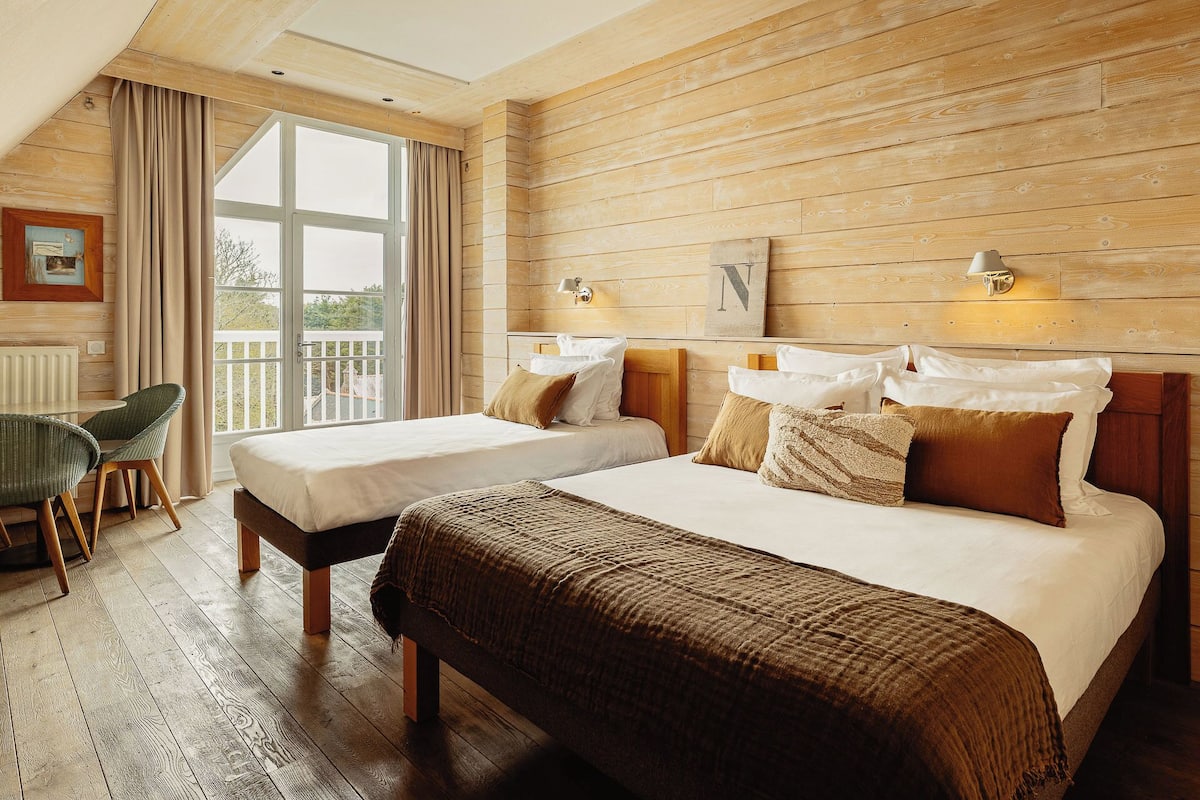 Une chambre d’hôtel avec deux lits, des murs en bois, une vue dégagée sur l’extérieur grâce à une grande fenêtre et un balcon.