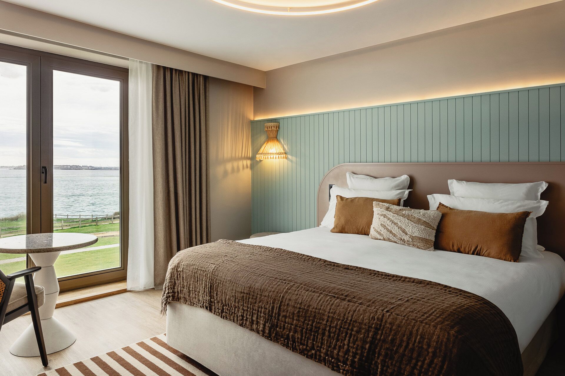 A hotel room with a large bed, a view of the water, a small table, and a lamp.